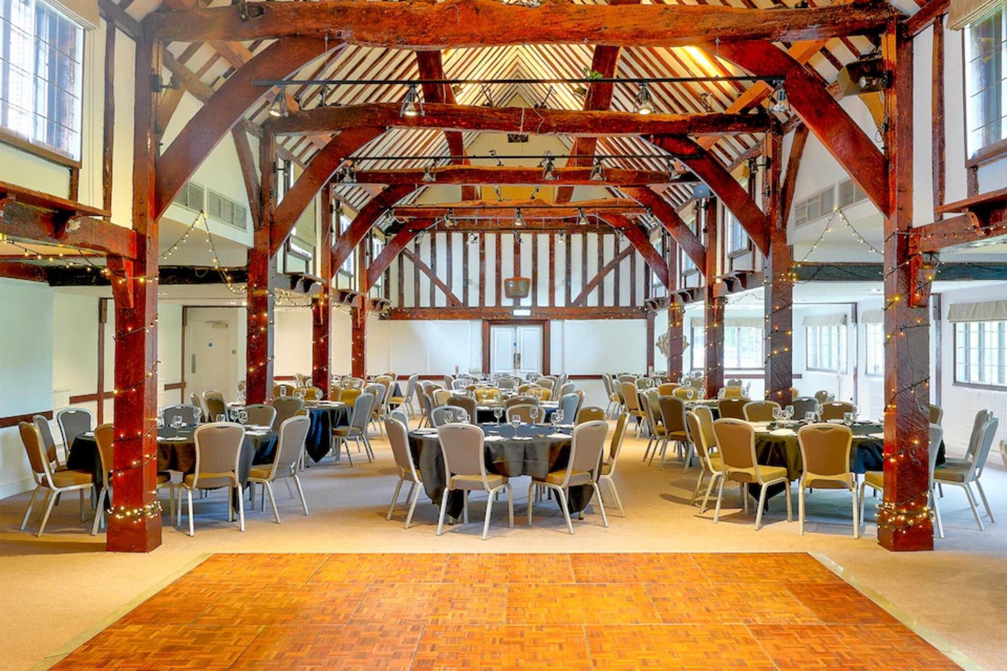 Een ruime zaal met een hoge plafond en zichtbare houten balken, voorzien van tafels en stoelen, geschikt voor evenementen.