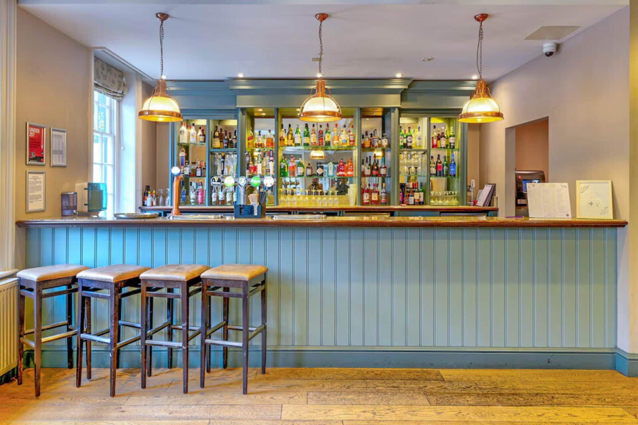 A bar area with wooden stools, a counter with a variety of bottles, and pendant lighting.