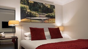 Habitación de hotel con cama, dos almohadas rojas, una colcha roja, un cuadro de un jardín y dos lámparas.
