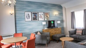 A hotel room with a round wooden table, orange chairs, a grey sofa, a TV, and wall art. - Sunday Walton Hall Retreat & Spa | Standard Double (Wellesbourne Hastings)