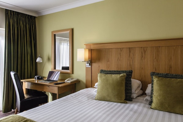 A hotel room with a bed, a desk, a chair, a lamp, a mirror, and a window with curtains. - Sunday Walton Hall Retreat & Spa | Standard Double (Wellesbourne Hastings)