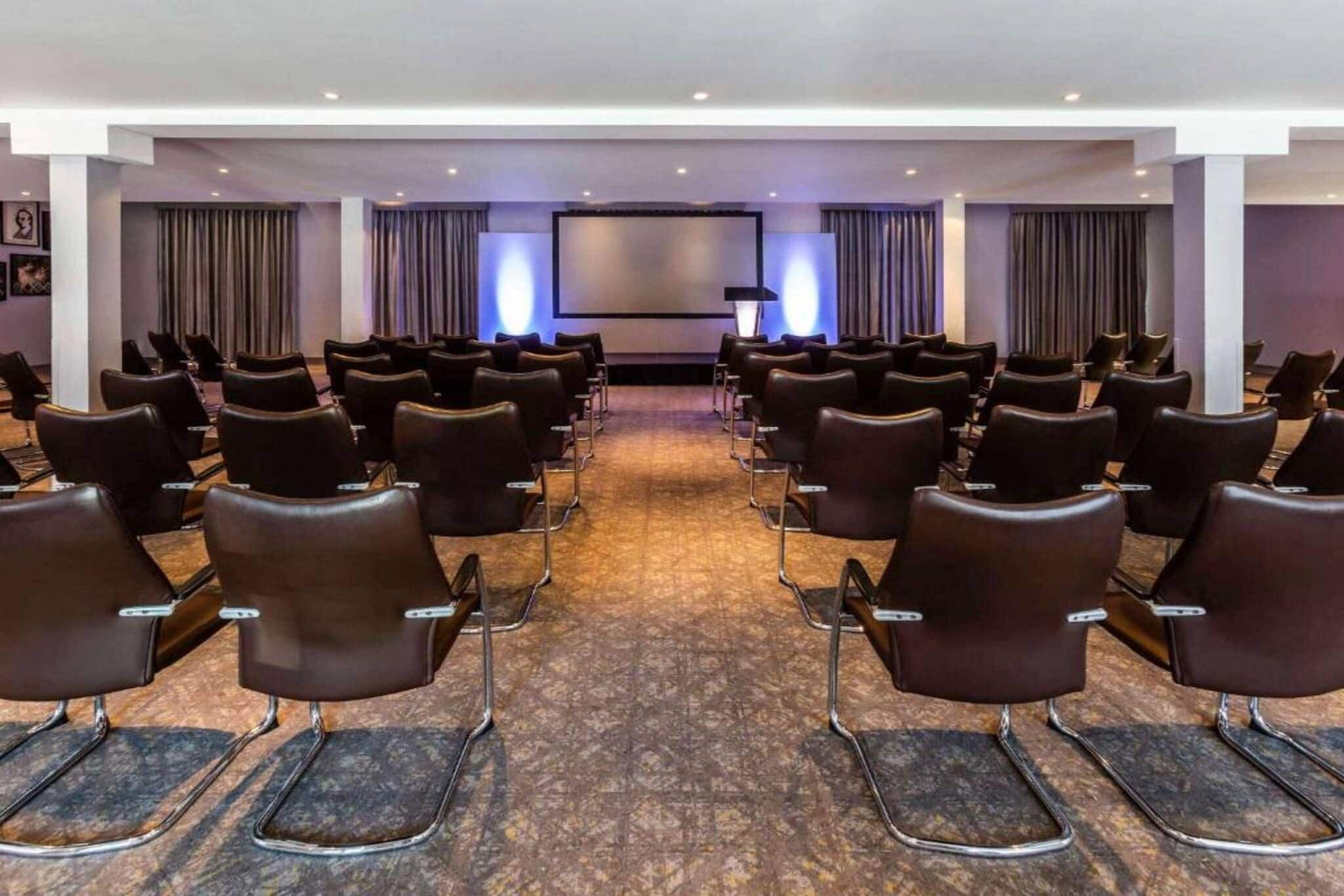 A conference room with rows of chairs facing a screen, carpeted floor, and curtains on the walls.