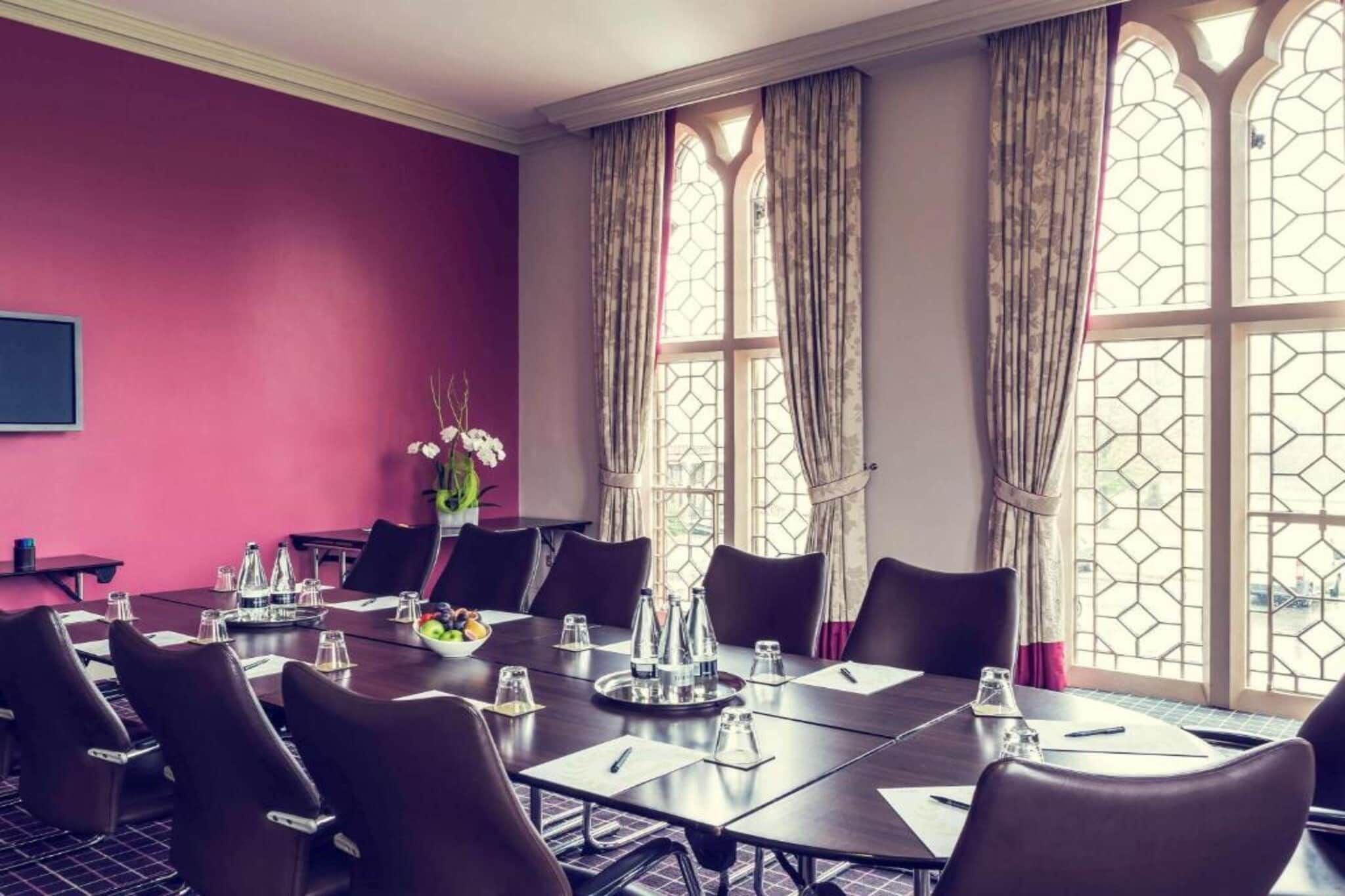 Sala de conferências com uma mesa comprida, cadeiras e uma grande janela com cortinas.