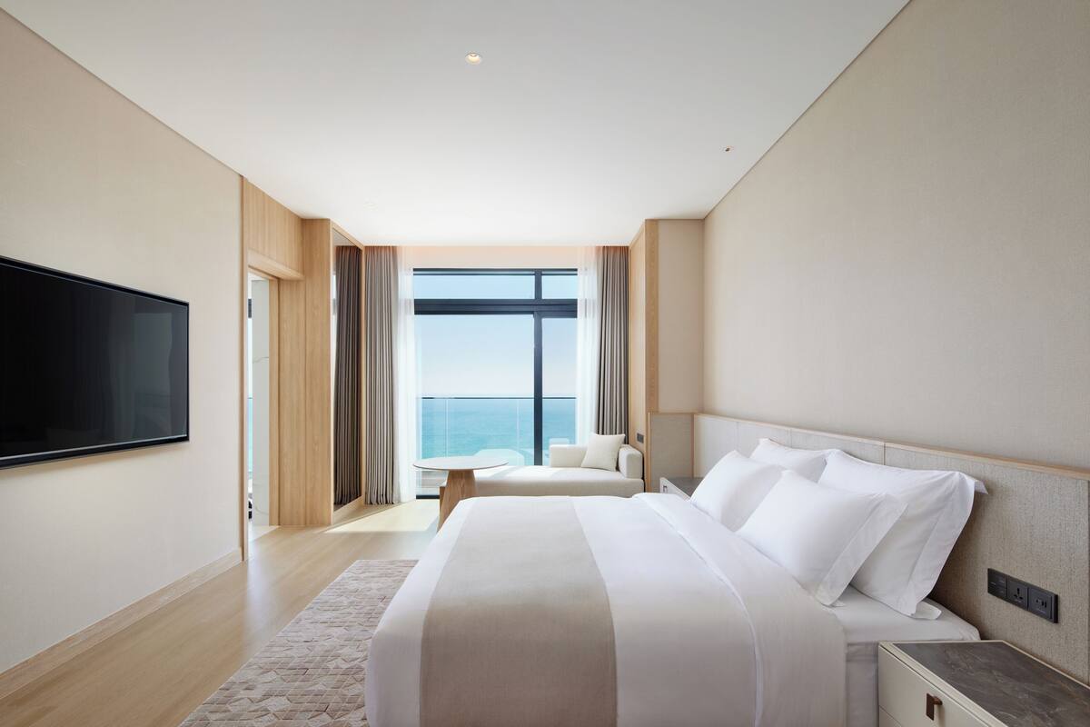A modern hotel room with a large bed, a flat-screen TV, a small table, and a view of the ocean through large windows.