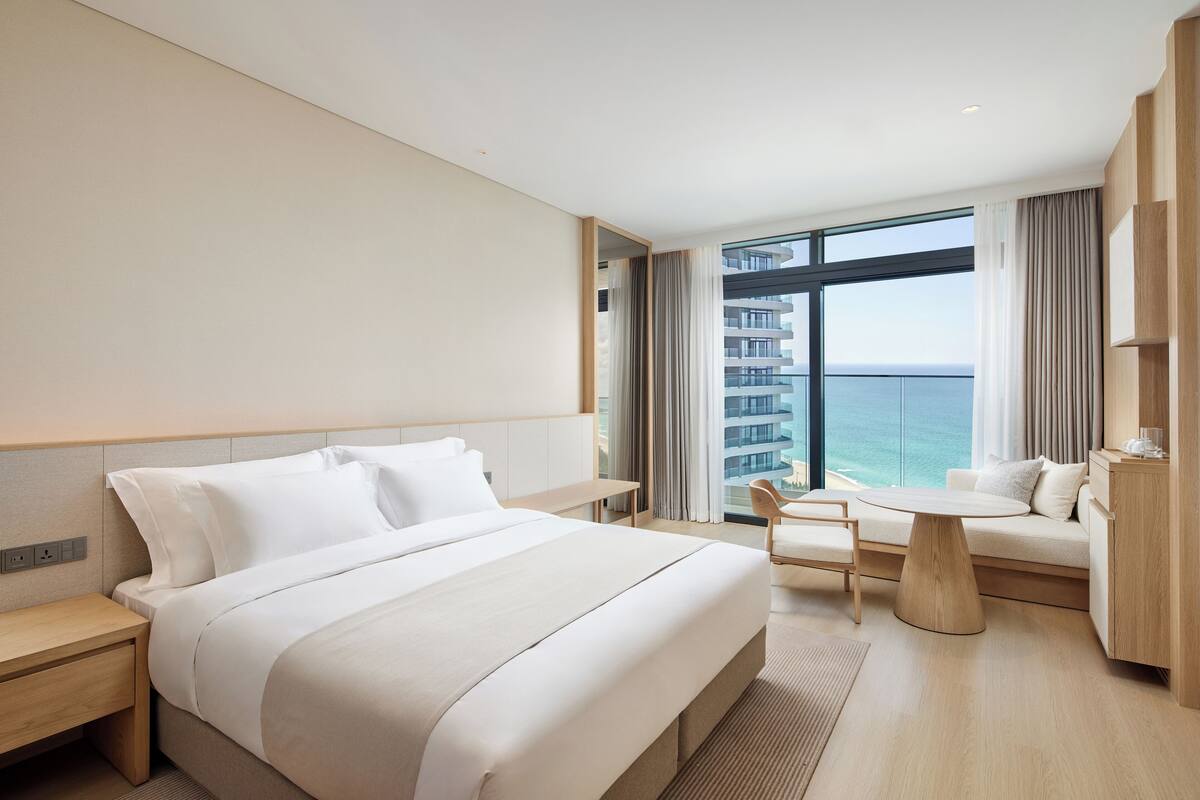A modern hotel room with a large bed, a seating area with a view of the ocean, and a balcony with a cityscape view.