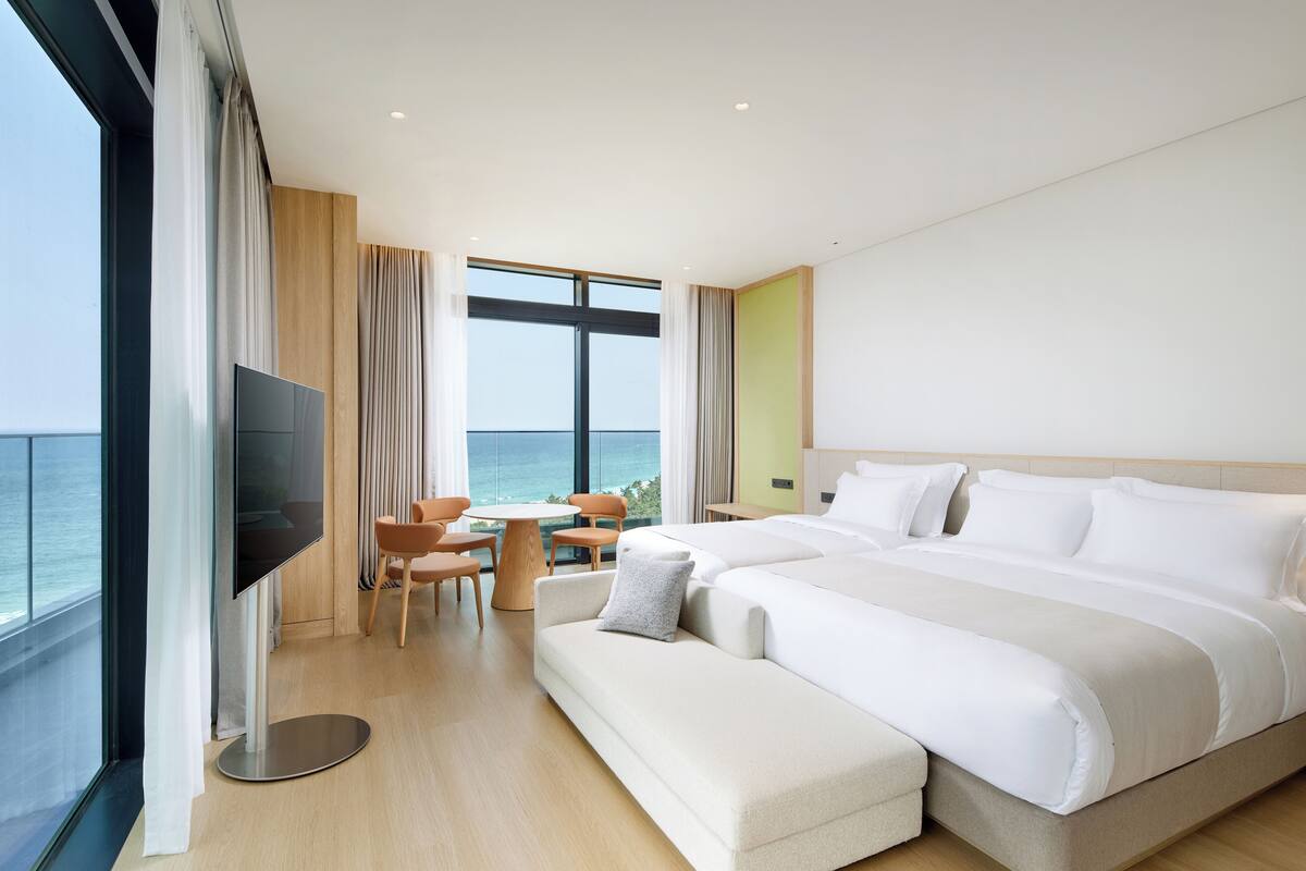 A modern hotel room with a large bed, a TV, a small table with chairs, and a view of the ocean.