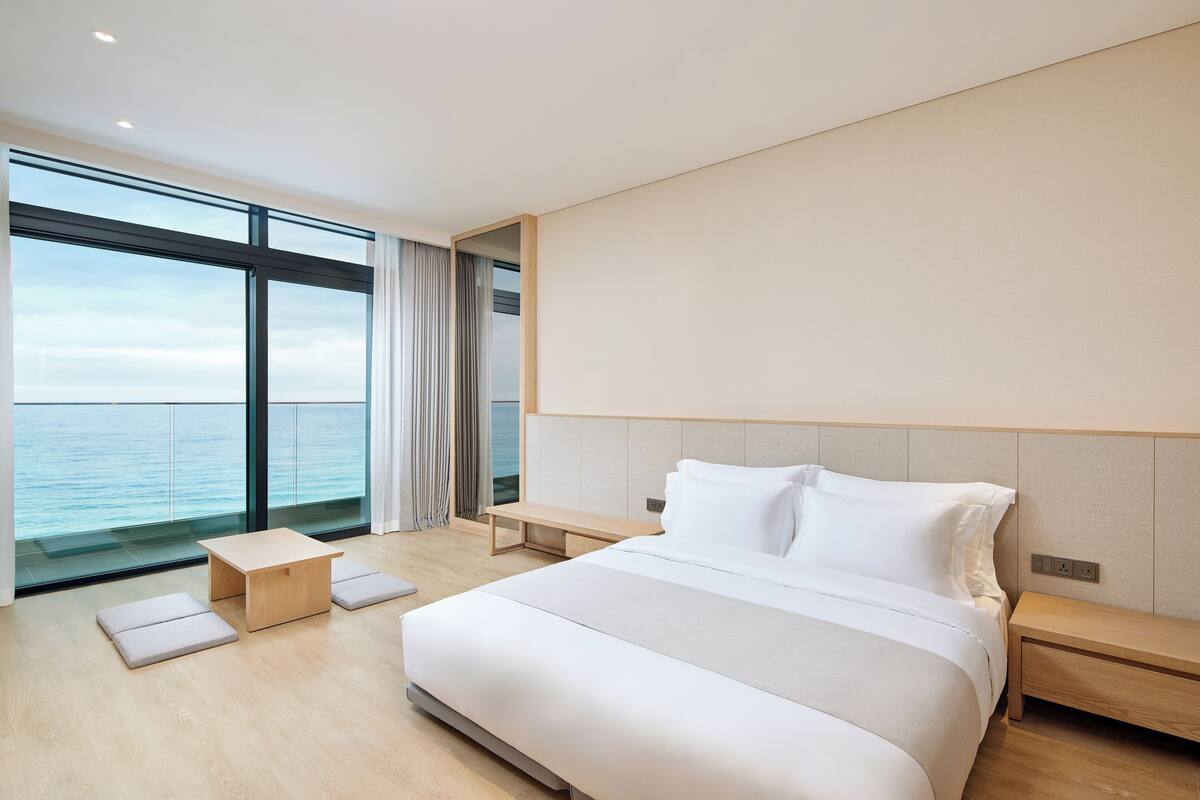 A modern hotel room with a large bed, a wooden nightstand, a small wooden table, and a bench.