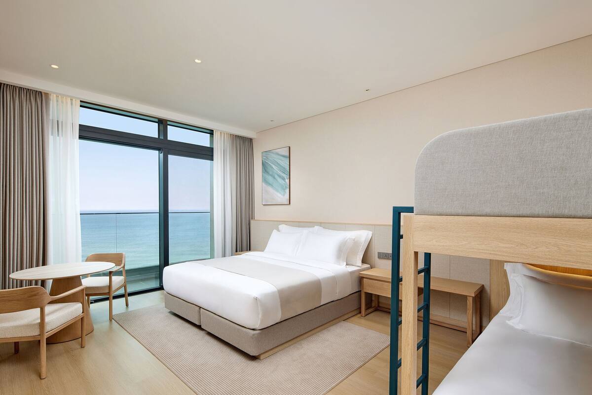 A modern hotel room with a bed, a desk, a chair, and a view of the sea.