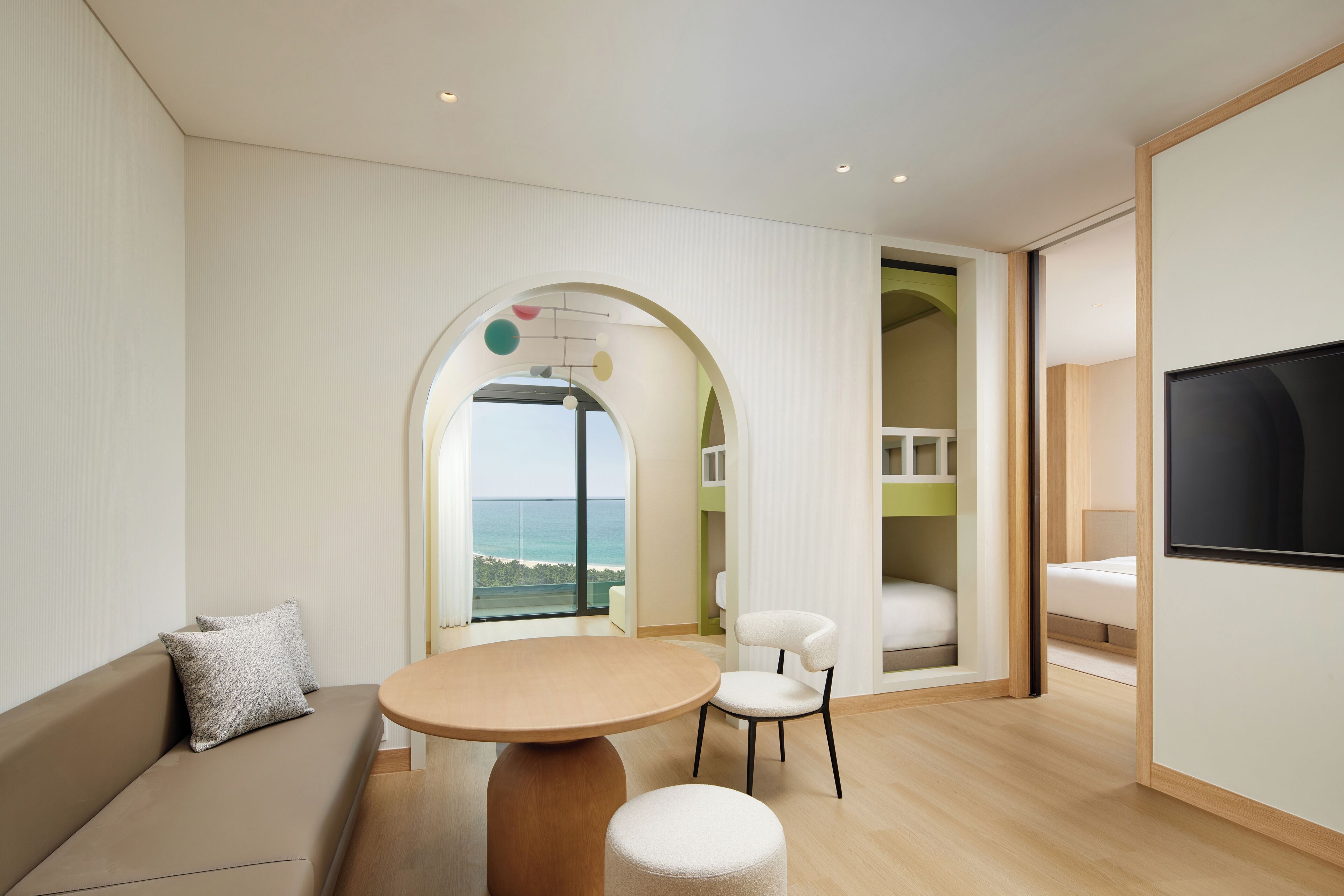 A modern hotel room with a round table, a sofa, a TV, and a view of the sea.