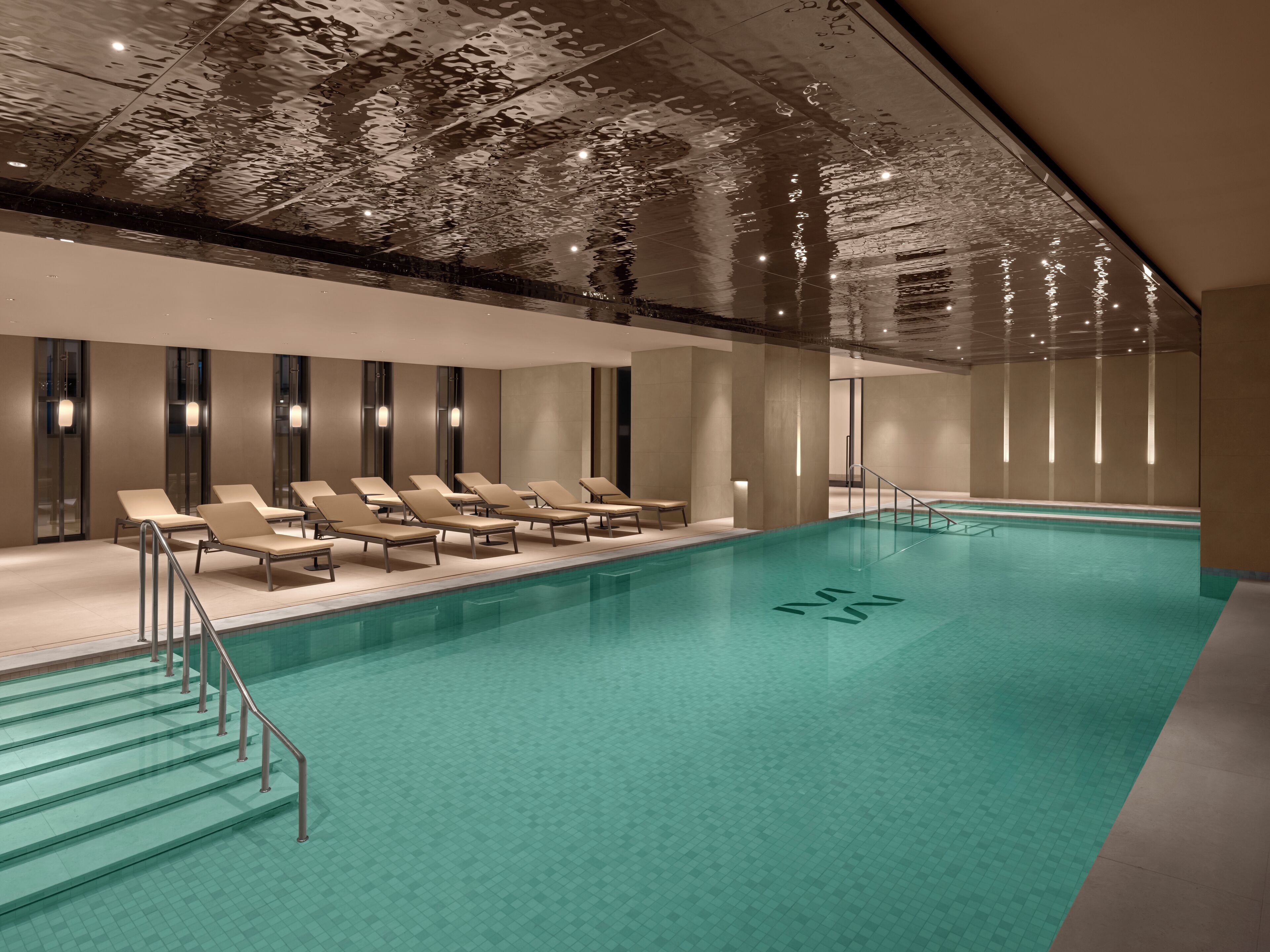 Una piscina interior moderna con agua cristalina, rodeada de sillas de descanso y una escalera que lleva a la piscina.