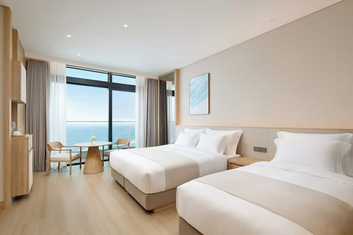 A hotel room with two beds, a dining table, a chair, a view of the ocean, and a large window.