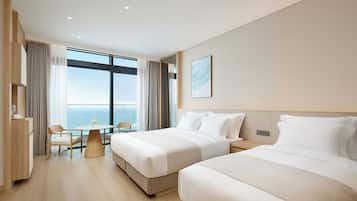 A hotel room with two beds, a dining table, a chair, a view of the ocean, and a large window.