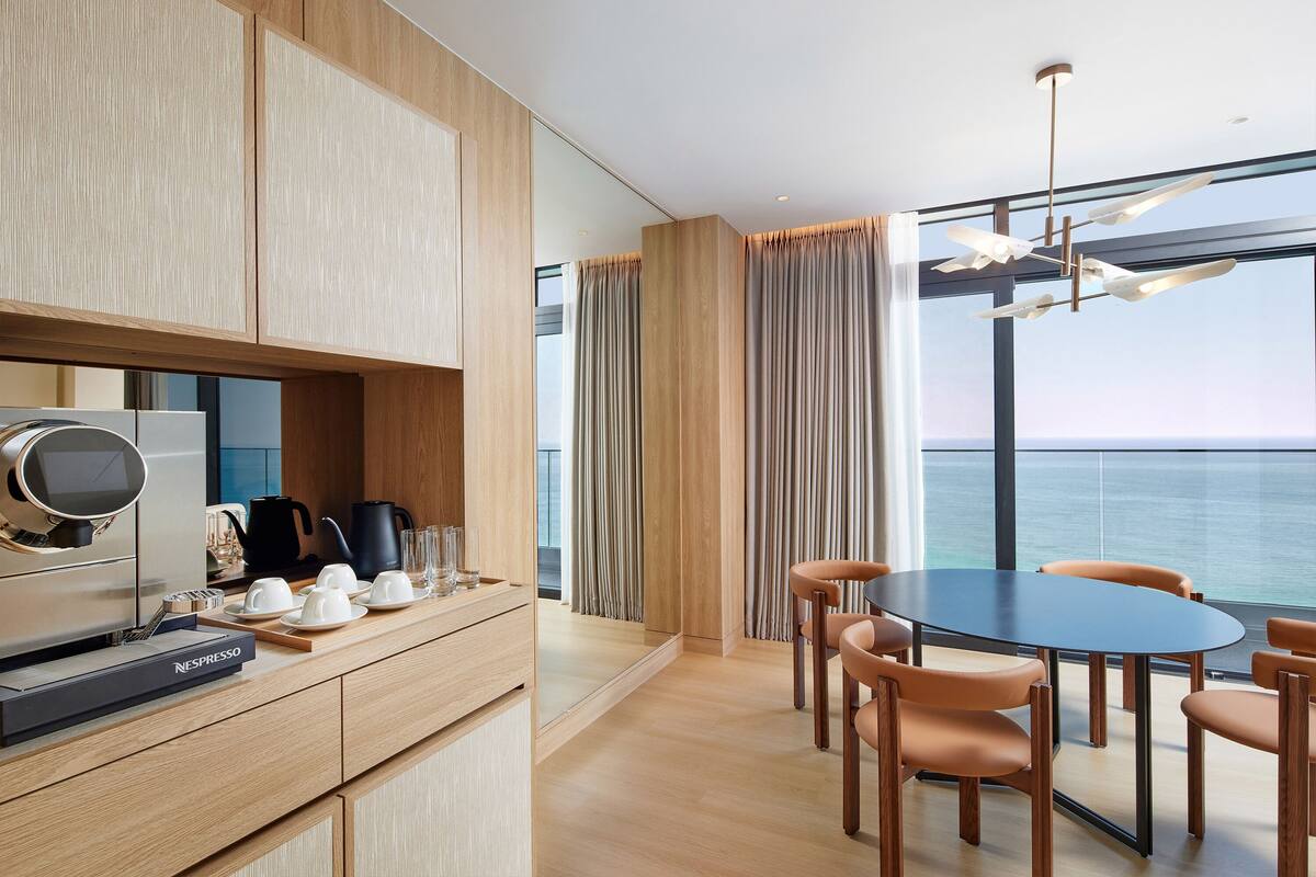 A modern kitchen with a coffee machine, a dining area with a round table and chairs, and a large window with a view of the sea.
