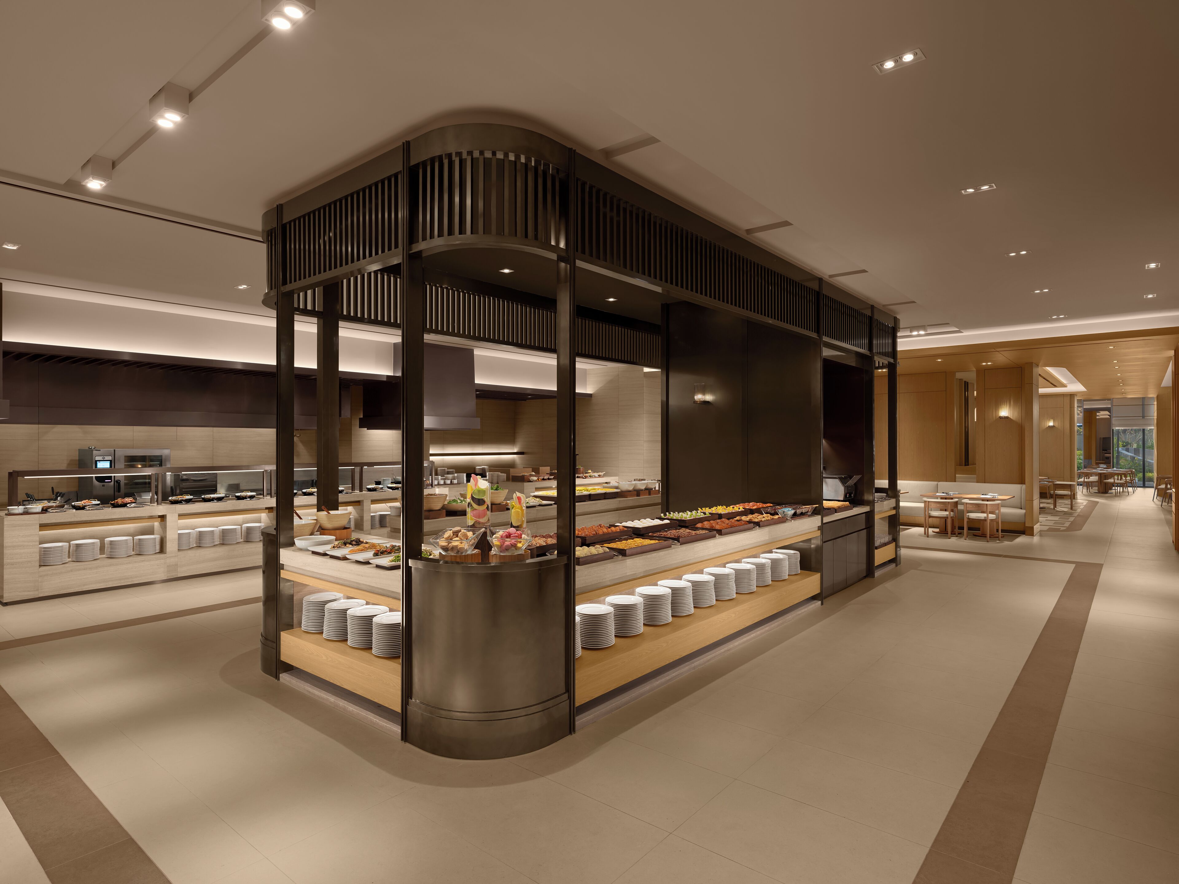 Un moderno vestíbulo de hotel con una zona de buffet que ofrece diferentes puestos de comida y un área de descanso con mesas y sillas.