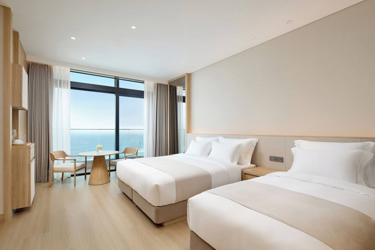 A modern hotel room with two beds, a dining table, and a view of the ocean.