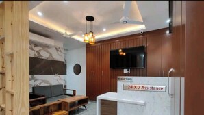 A reception desk with a sign indicating 24x7 assistance, a television mounted on the wall, and a wooden reception desk with a marble-like design. - Hotel Blue Shine  (Noida)