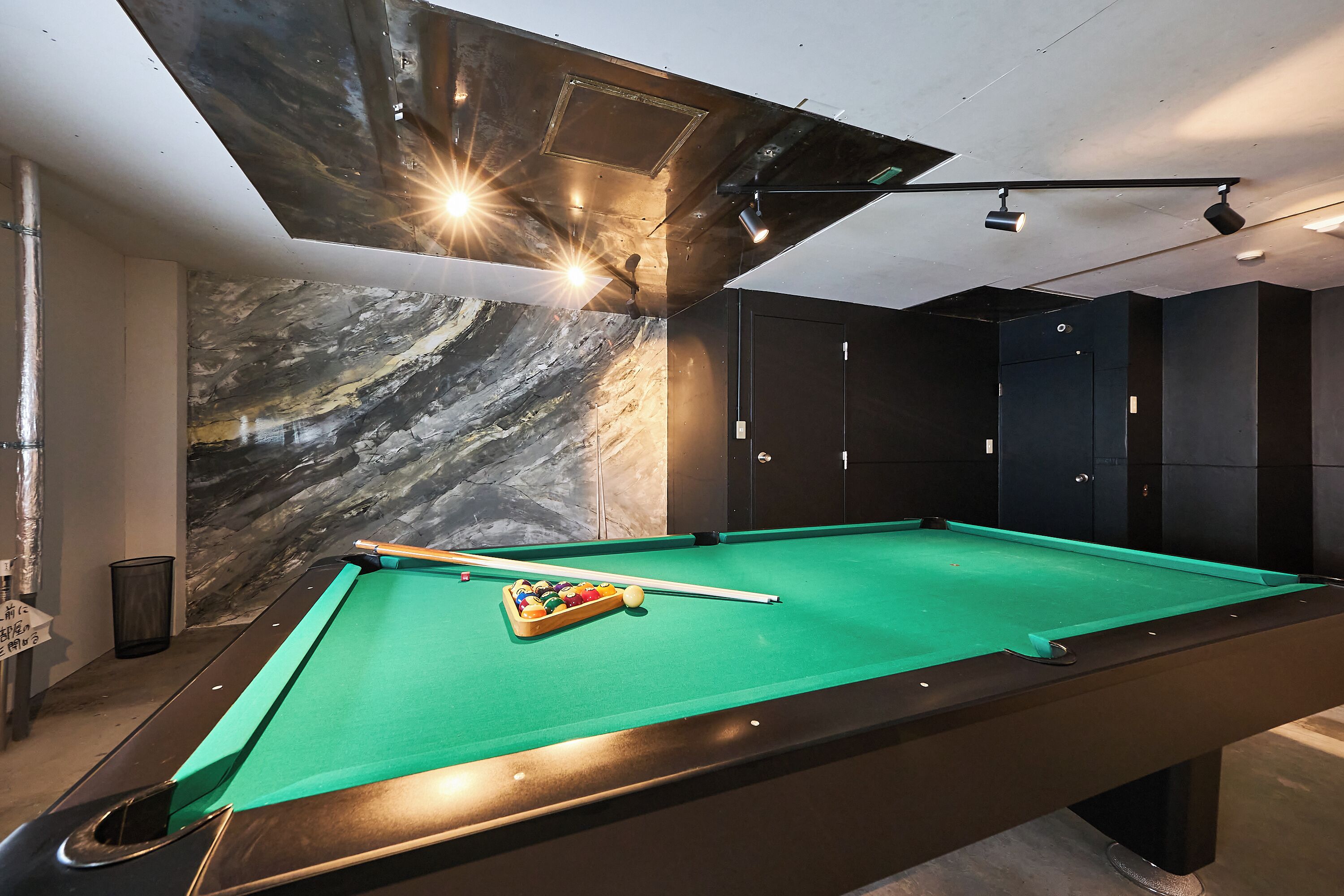 Une table de billard recouverte d’un drap vert, entourée d’armoires aux couleurs sombres et adossée à un mur décoré d’un motif imitant le marbre.