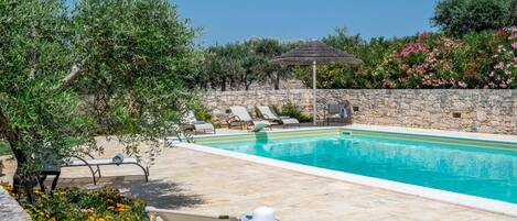Un área junto a la piscina con sillas de descanso, un muro de piedra y una sombrilla de paja.