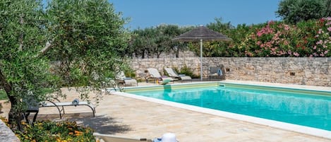 Un espace aménagé au bord de la piscine, avec des chaises longues, un mur en pierre et un parasol en chaume.