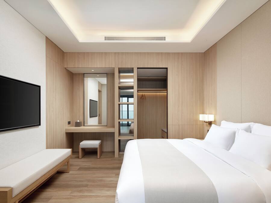 A modern hotel room with a large bed, a flat-screen TV, a desk with a chair, a bench, and a walk-in closet.