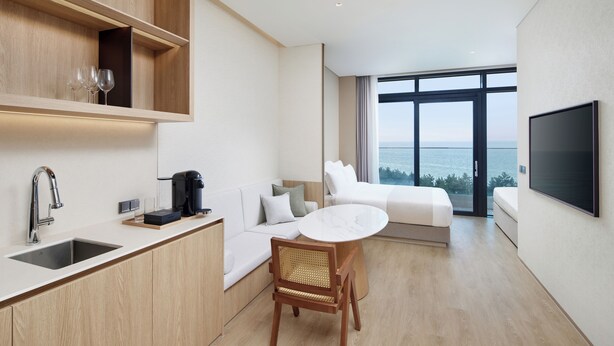 A modern hotel room with a kitchen area, a dining table, a sofa, and a large window offering a view of the sea. - Shillamonogram Gangneung Residence (Gangneung)