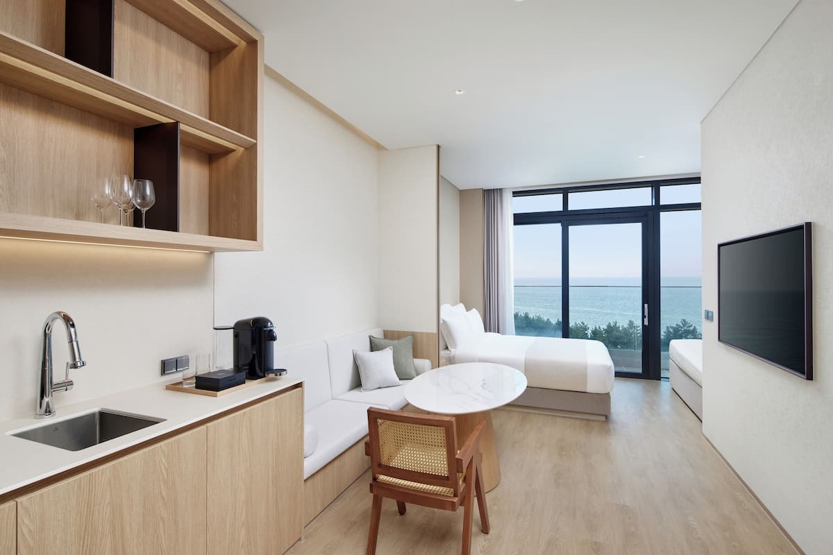 A modern hotel room with a kitchen area, a dining table, a sofa, and a large window offering a view of the sea.