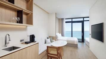 A modern hotel room with a kitchenette, a dining area, a sofa, a bed, and a large window offering a view of the sea.