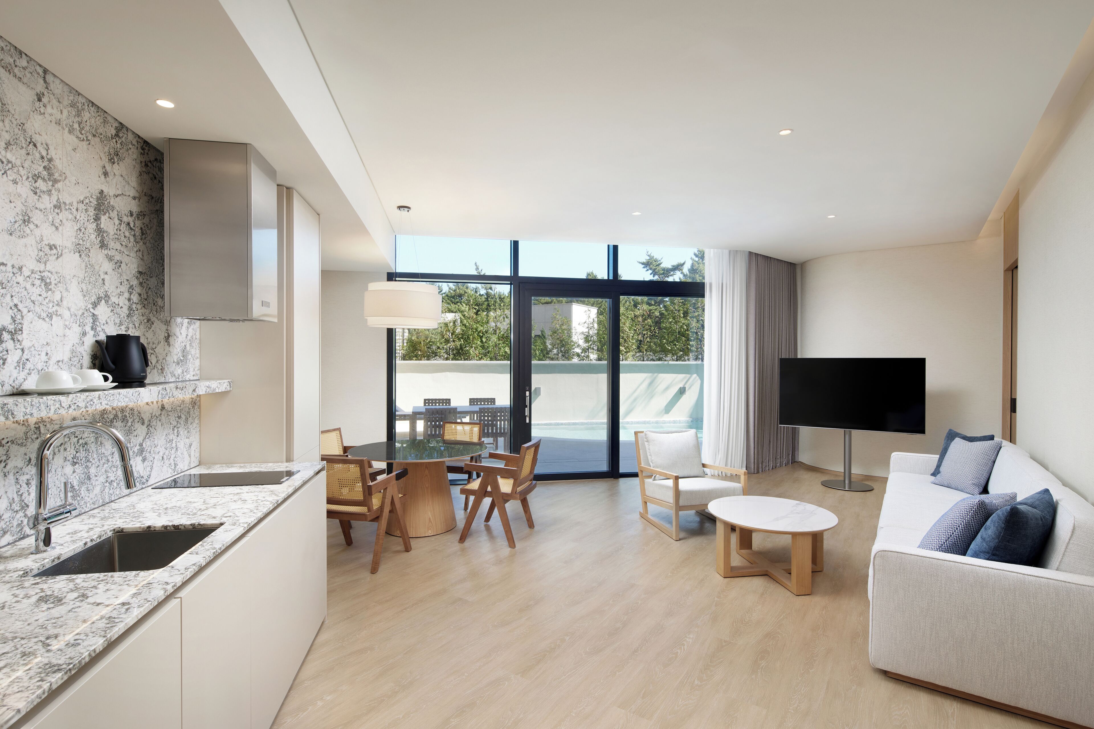 Cocina moderna con encimera de mármol y electrodomésticos de acero inoxidable, comedor con mesa y sillas redondas, sala de estar con sofá y televisor de pantalla plana, y ventanas amplias con vistas al exterior.
