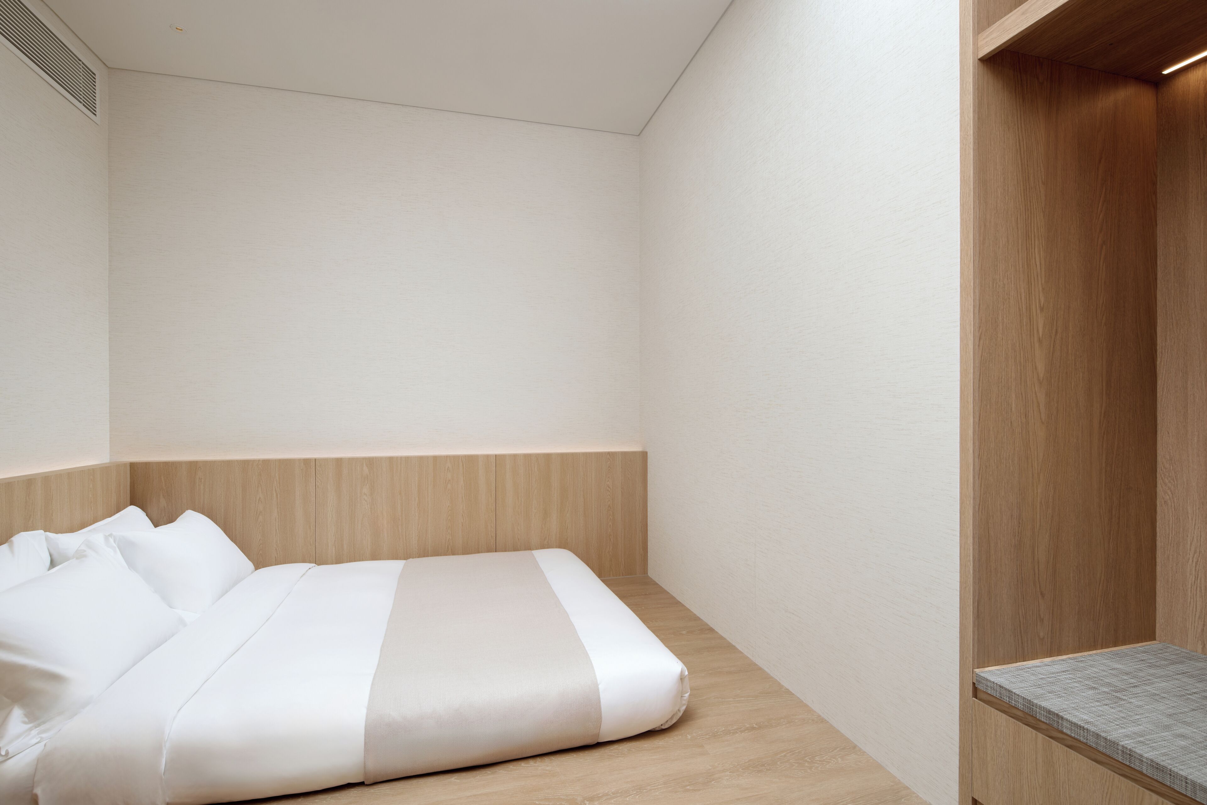 A minimalist hotel room with a bed, a wooden bench, and a small seating area.