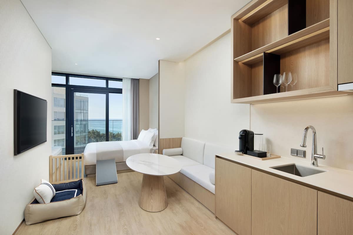 Ein modernes Hotelzimmer mit Bett, Sofa, Esstisch und Küchenzeile inklusive Spüle, sowie ein großes Fenster mit Aussicht.