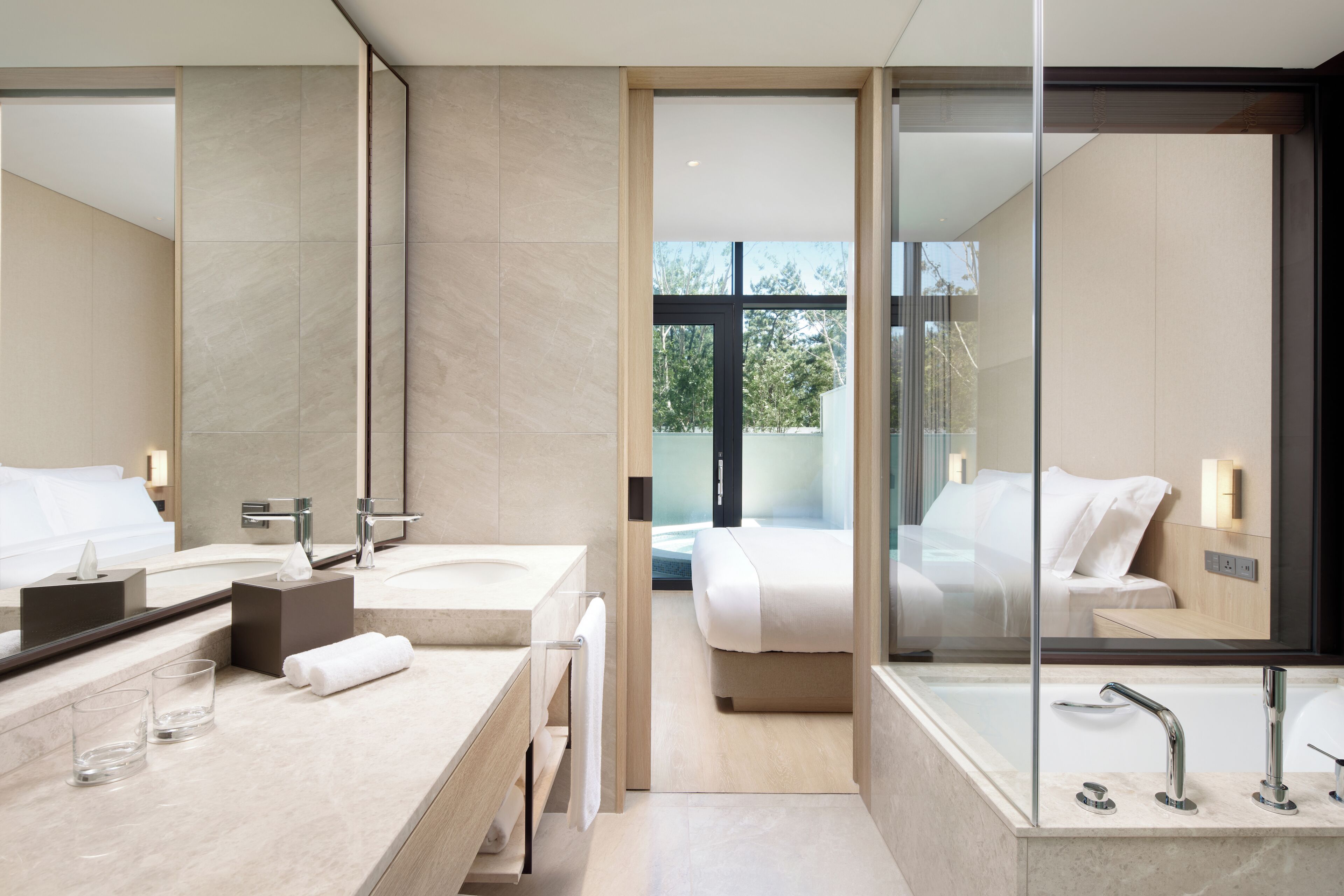 Ein modernes Hotelbad mit einem großen Spiegel, zwei Waschbecken, einer Badewanne und einem sichtbaren Schlafzimmer mit Bett und Fenster.
