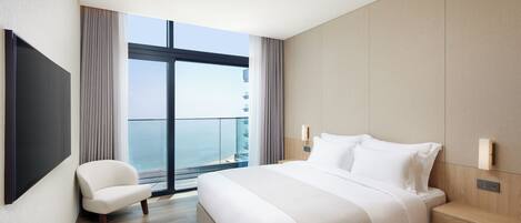 A modern hotel room with a large bed, a chair, a TV mounted on the wall, and a view of the sea through a large window.