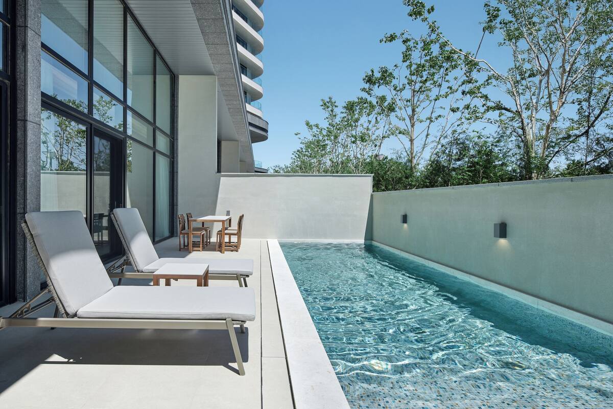 A modern outdoor pool area with lounge chairs and a table, surrounded by a high wall and trees.