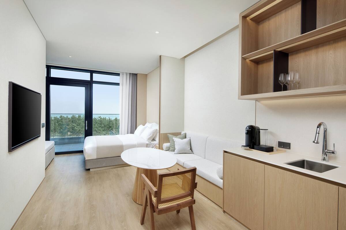 A modern hotel room with a bed, a sofa, a dining table, a coffee maker, and a sink.