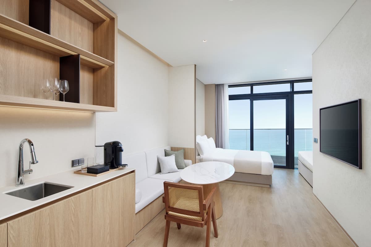 A modern hotel room with a kitchen area, a dining table, a sofa, a bed, and a TV.