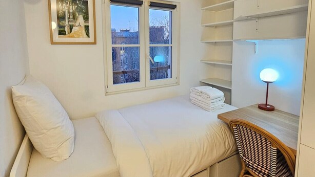 A compact bedroom with a bed, a desk, and a chair. There are two windows with blinds, a wall-mounted shelf, and a framed picture. - Emily in Paris - Panthéon - Luxembourg - Studio (Paris)