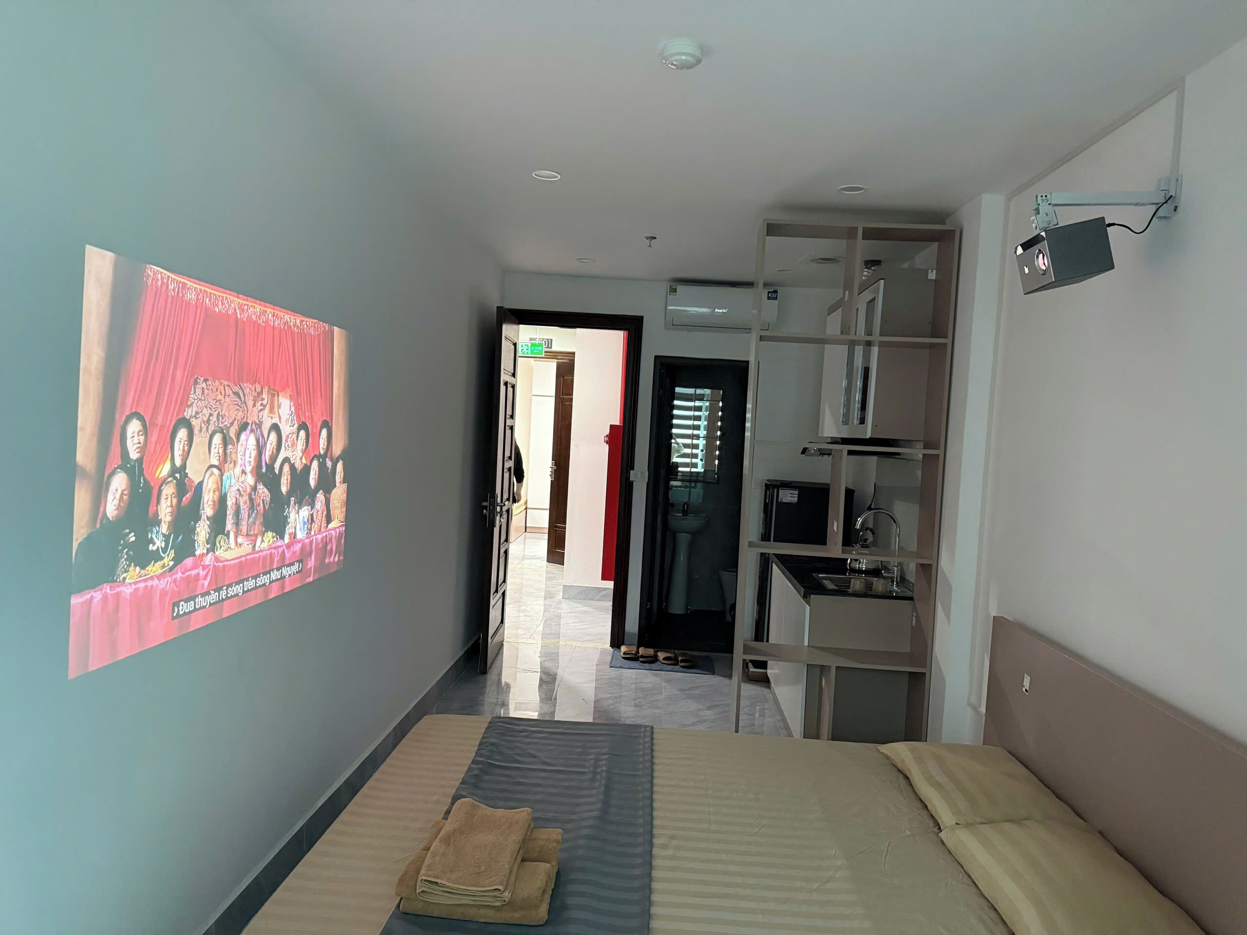 Quarto de hotel com uma cama, uma televisão mostrando uma foto de grupo, um guarda-roupa e um banheiro visível por uma porta aberta.