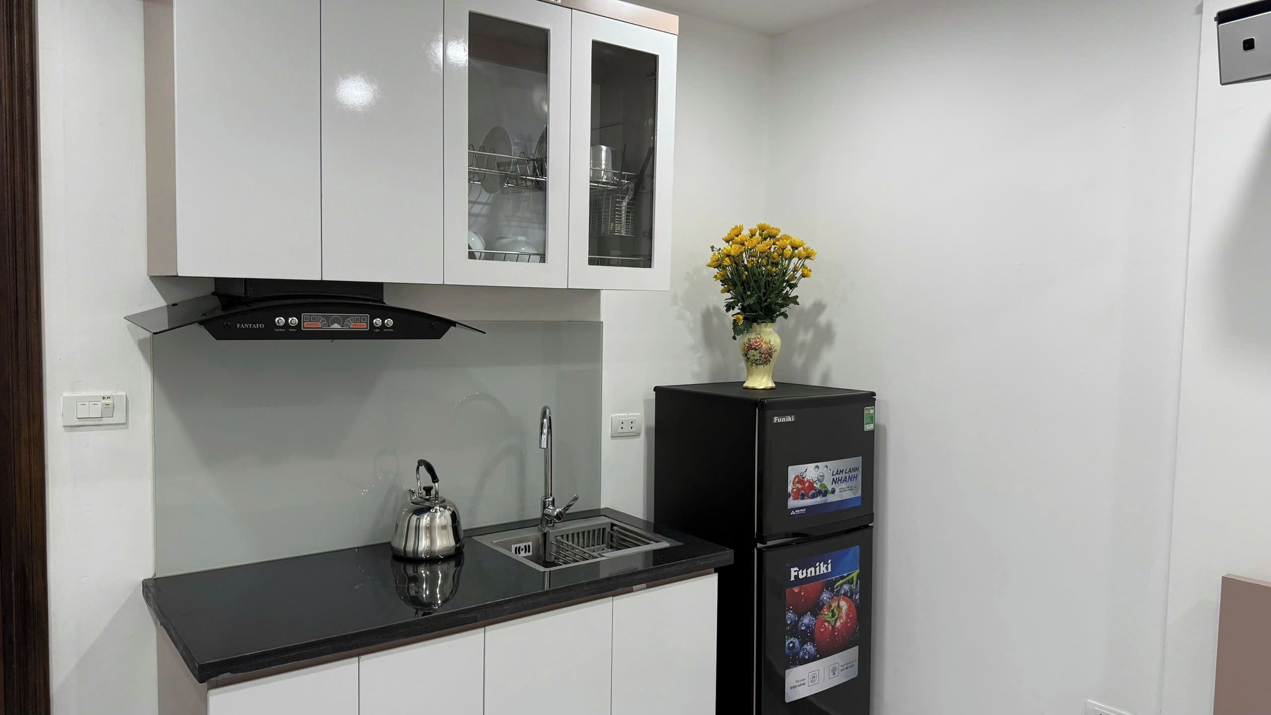 A compact kitchen with a black countertop, a kettle, a sink, and a refrigerator.