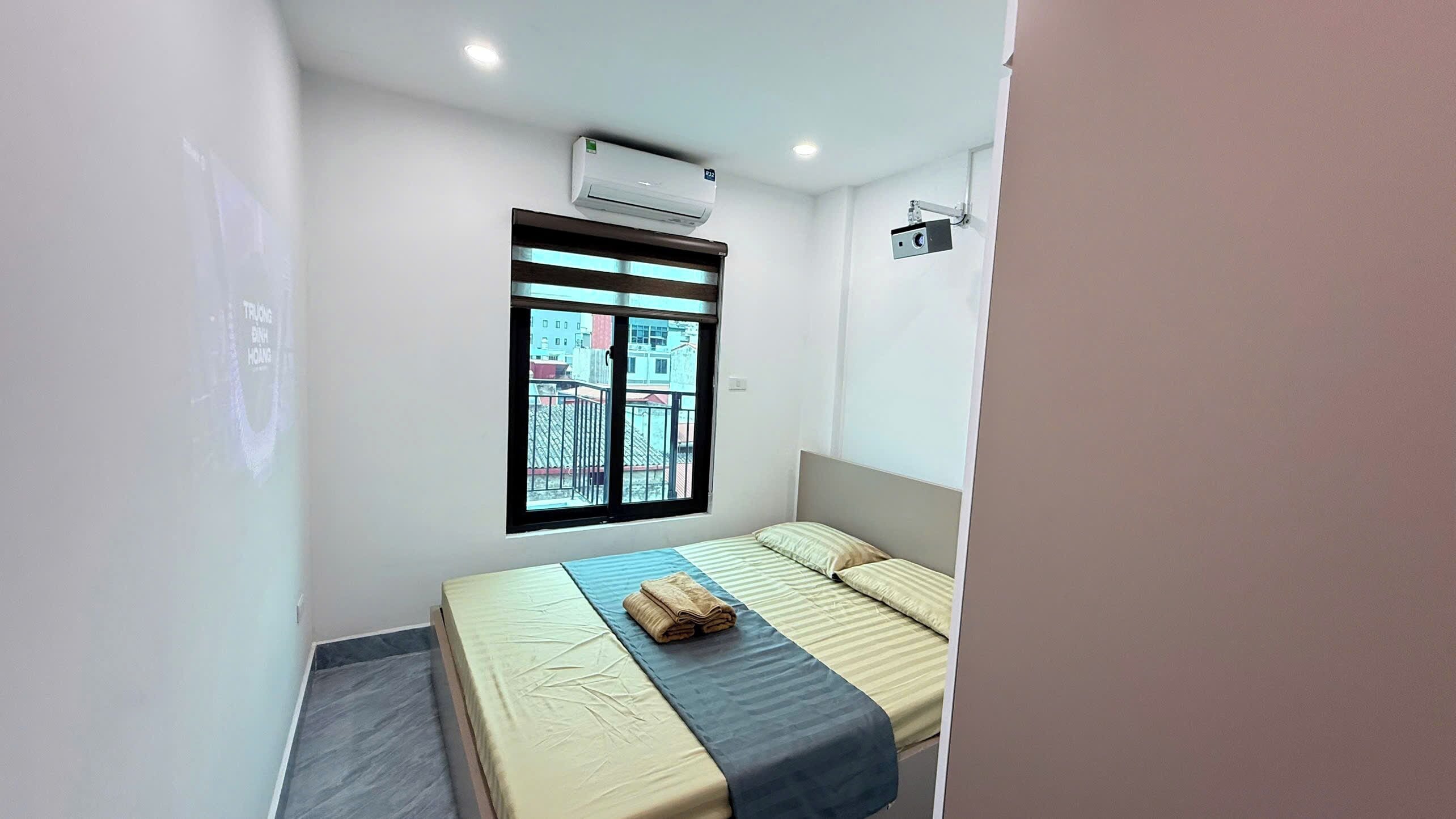 A small, modern bedroom with a single bed, a window with a view of buildings, an air conditioning unit, and a security camera.