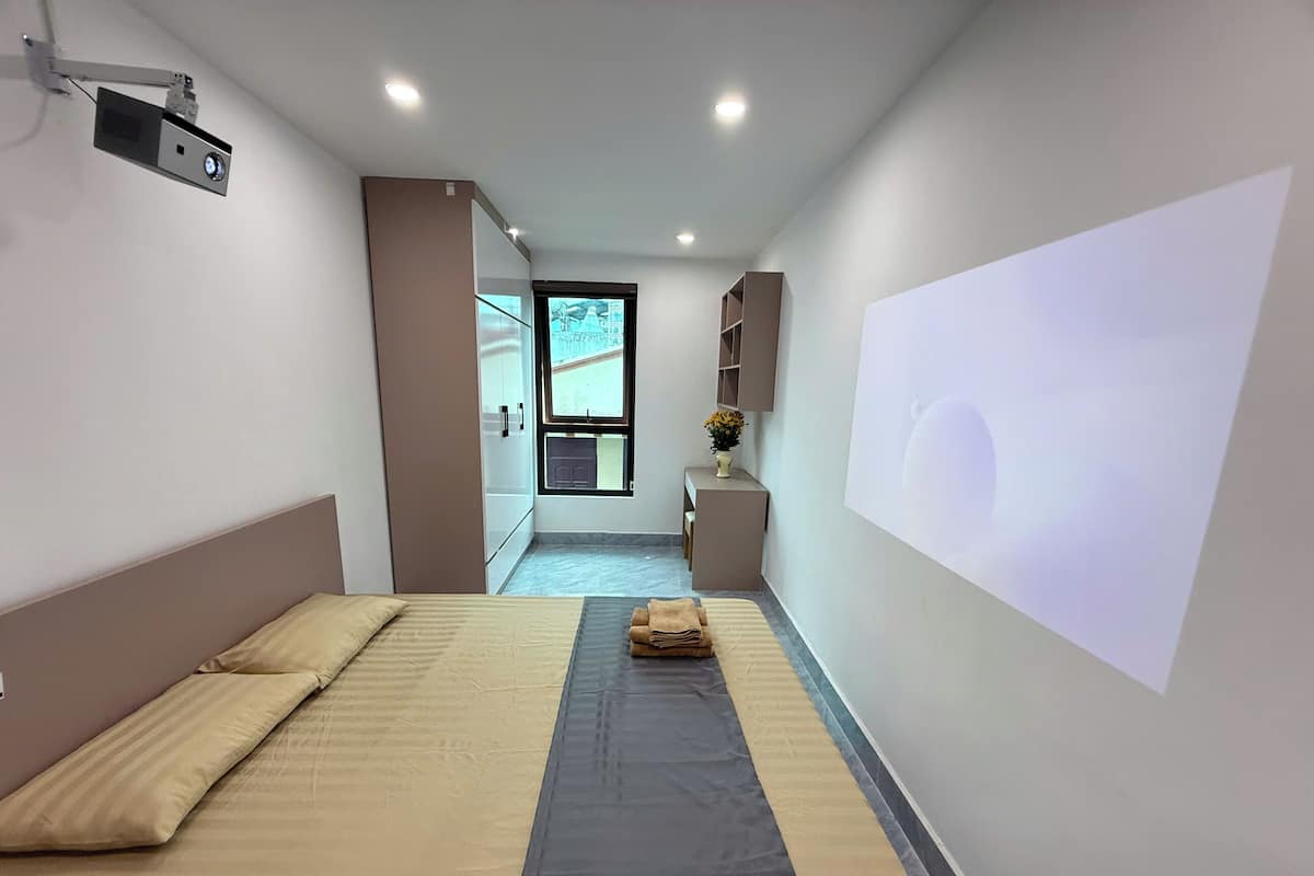 Habitación de hotel con cama, un pequeño escritorio, una silla, una ventana y un proyector montado en la pared.