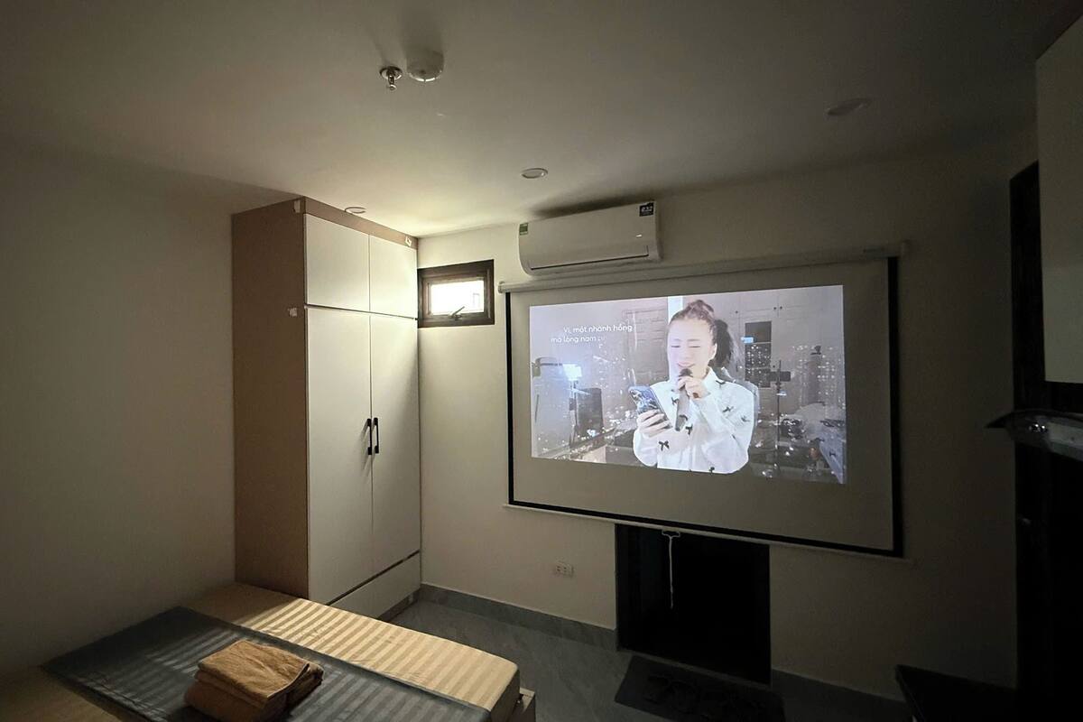 Una habitación con una pantalla grande que muestra una persona, una cama, un armario y una ventana pequeña.