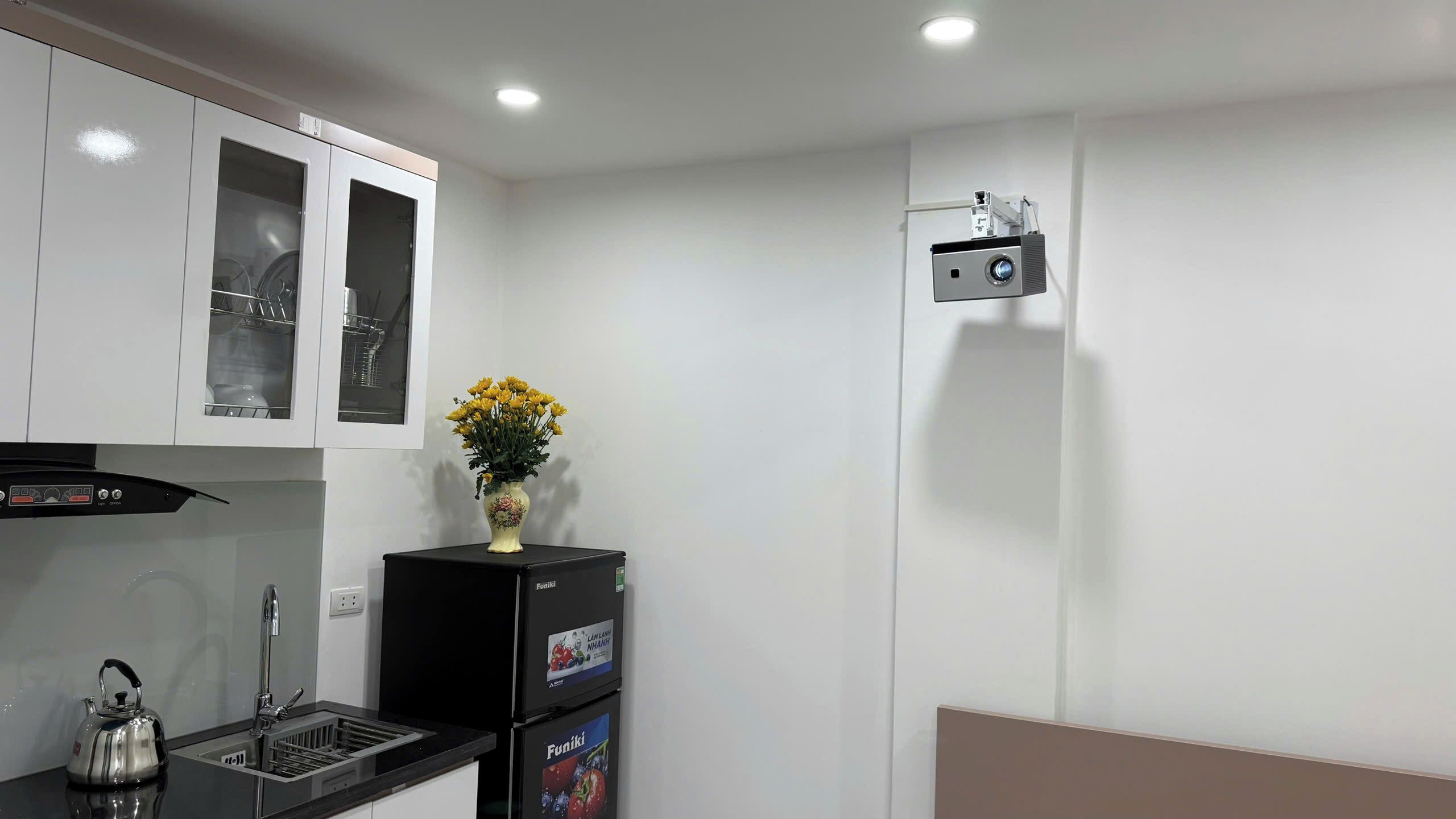 A compact kitchen with white cabinets, a black refrigerator, a kettle, and a small decorative vase with yellow flowers.