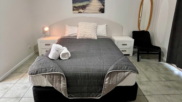 A bedroom with a bed, a nightstand, a chair, a mirror, and a painting of a beach. - Agnes palms motel (Agnes Water)