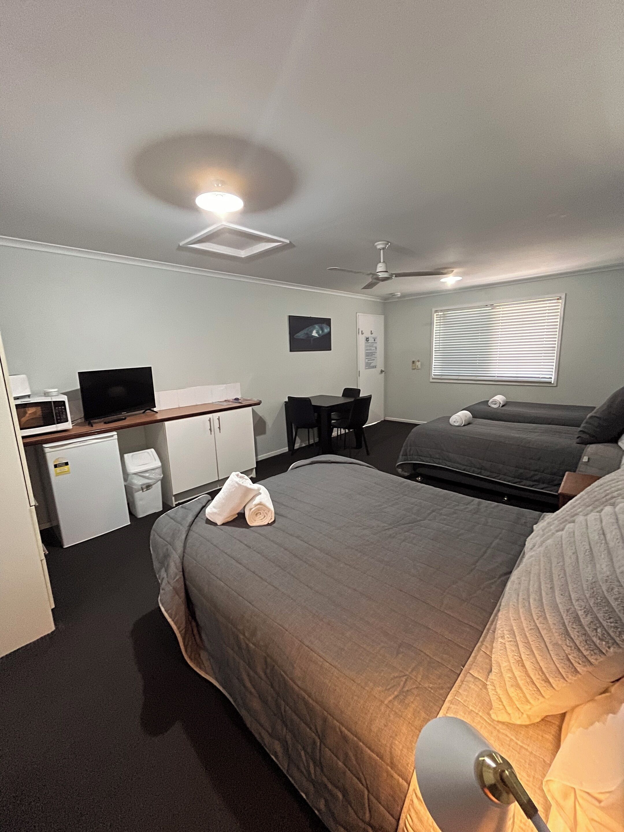 A compact hotel room with a bed, a desk with a computer, a microwave, and a ceiling fan.