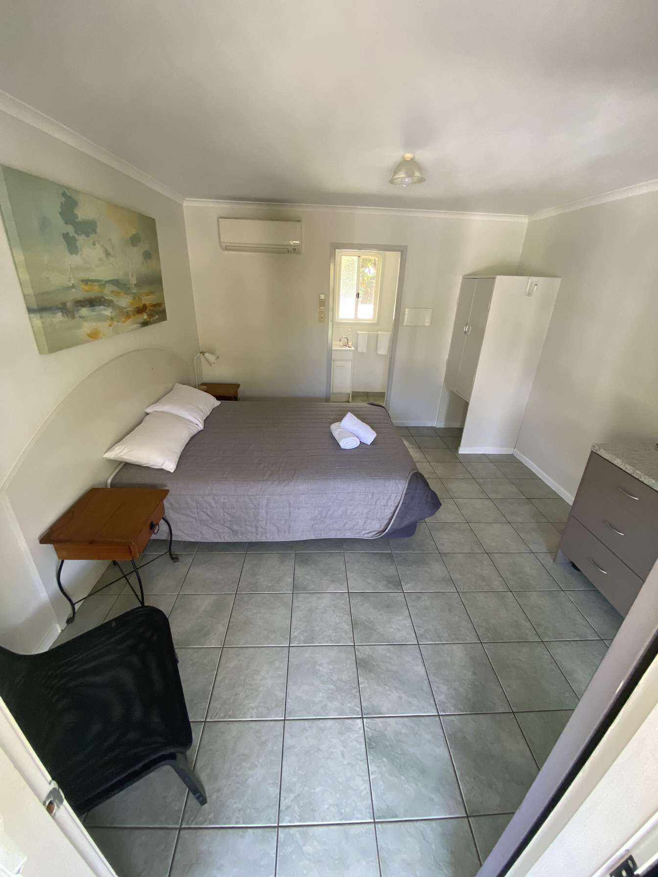 Een slaapkamer met een bed, een klein tafeltje, een stoel, een kledingkast en een schilderij aan de muur.