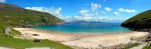 Ein Küstenstrand mit einem kleinen Gebäude, klarem, blauem Wasser und grünen Hügeln.