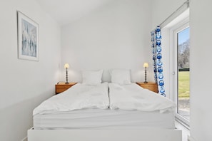A bedroom with a bed, two bedside tables, a lamp, a window with curtains, and a framed picture on the wall.