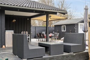 A covered outdoor seating area with a table and chairs, a pergola, and a small building in the background.