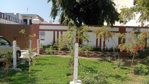 A building with a brick facade, a white fence, and a grassy area with small trees. - 2-bedroom villa in welcoming Faridabad with WiFi, AC, badminton court within. (Faridabad)