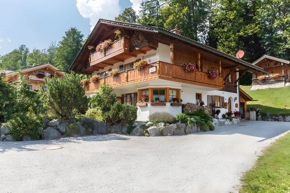 A traditional Alpine-style building with multiple balconies, a stone wall, and a gravel driveway.
